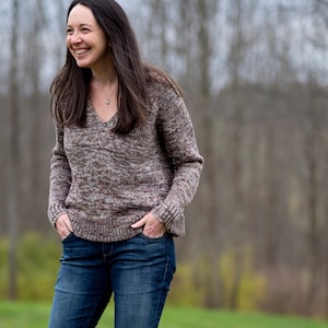 Może przedstawiać: Kobieta w brązowym i białym wzorzystym swetrze z dekoltem w kształcie litery V, niebieskich dżinsach i brązowych butach. Stoi na trawiastym polu z drzewami w tle.