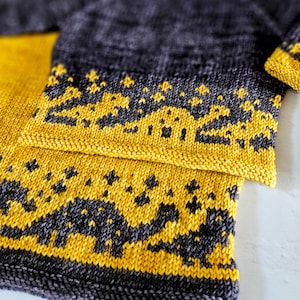 Op de afbeelding: Close-up van een gebreide stof met een geel en grijs patroon. De stof is dubbelgevouwen, waardoor het patroon aan beide kanten zichtbaar is.