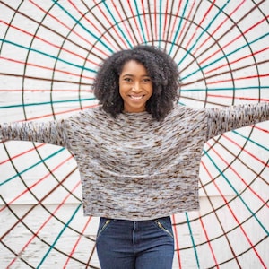 Puede incluir: Una mujer con el pelo oscuro y rizado está de pie frente a una pared blanca con un patrón geométrico colorido. Lleva un suéter gris y marrón y pantalones vaqueros azules. Está sonriendo y mirando a la cámara.
