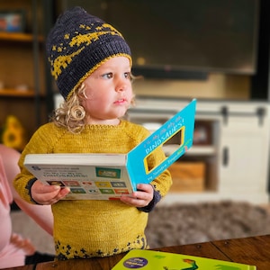 May include: A young child wearing a blue and yellow knit hat and a yellow sweater is reading a book titled "Oh My Oh My Dinosaurs!" The book has a blue cover with a yellow window.