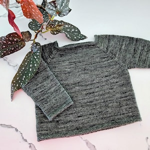 Könnte beinhalten: Ein grauer Strickpullover mit langen Ärmeln. Der Pullover ist aus strukturiertem Garn gefertigt und hat einen gerippten Saum und Bündchen. Der Pullover wird auf einer weißen Oberfläche mit grünen und roten Blättern präsentiert.