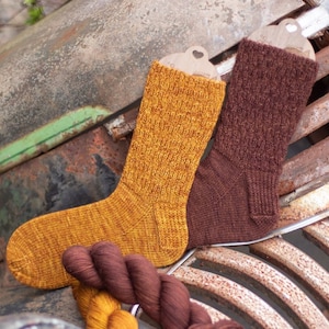May include: Two pairs of knitted socks, one in a brown color and one in a yellow and orange color. The socks are sitting on a rusty metal surface.