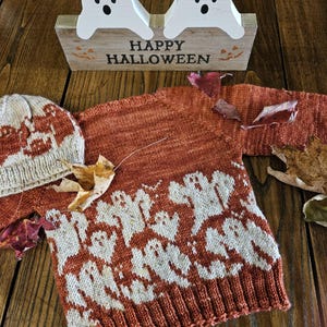 May include: A rust-colored knitted sweater and matching hat with a white ghost pattern. A wooden sign with two white ghost figures and the words "HAPPY HALLOWEEN" is in the background. Autumn leaves are scattered around the sweater.