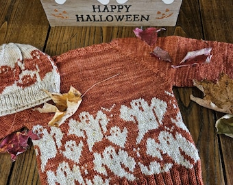 May include: A rust-colored knitted sweater and matching hat with a white ghost pattern. A wooden sign with two white ghost figures and the words "HAPPY HALLOWEEN" is in the background. Autumn leaves are scattered around the sweater.
