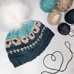 May include: A knitted winter hat with a teal and dark teal color scheme, adorned with a white faux fur pom-pom. The hat features a geometric pattern in beige and black. Several balls of yarn in various colors are arranged around the hat.