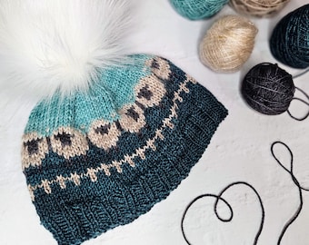 May include: A knitted winter hat with a teal and dark teal color scheme, adorned with a white faux fur pom-pom. The hat features a geometric pattern in beige and black. Several balls of yarn in various colors are arranged around the hat.
