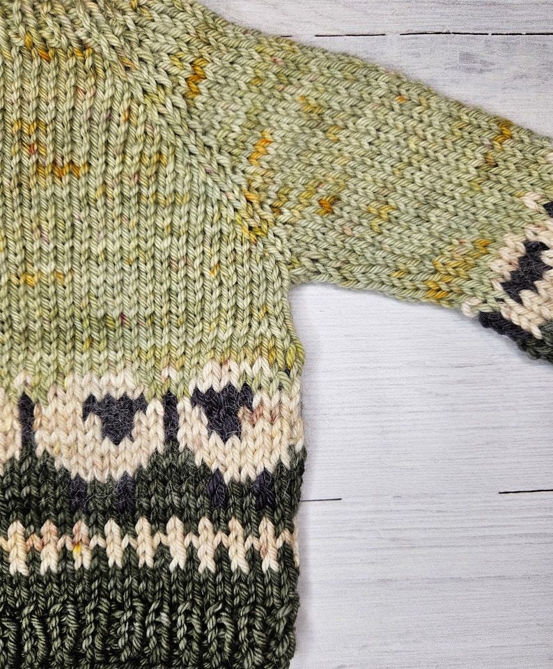 May include: A green knitted sweater with a pattern of white sheep and black accents. The sweater has a textured knit and a subtle yellow hue.