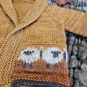 May include: A brown knitted cardigan with a shawl collar and a white and brown sheep pattern on the bottom. The cardigan is laying on a gray and white fuzzy surface.