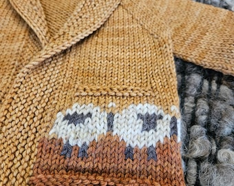 May include: A brown knitted cardigan with a shawl collar and a white and brown sheep pattern on the bottom. The cardigan is laying on a gray and white fuzzy surface.