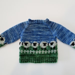 May include: A blue and green knitted sweater with a pattern of white sheep and black fences. The sweater has long sleeves and a crew neck.
