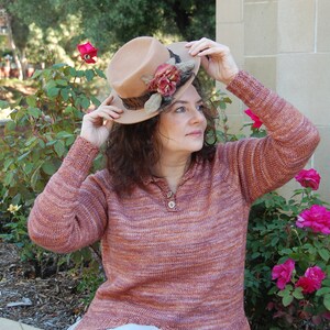 May include: A tan fedora hat with floral and leaf embellishments. The hat is being held up. The person is wearing a knit sweater in shades of orange and pink, and a light blue tulle skirt. The background includes rose bushes and a stone wall.
