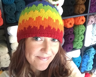 May include: A woman wearing a rainbow knit hat with a colorful yarn ombre design. The hat is made with a chunky yarn and has a slouchy fit. The woman is standing in front of a wall of yarn balls.