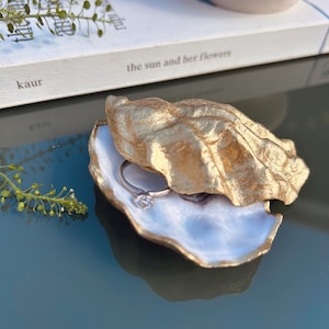 May include: A gold-painted oyster shell ring dish with a silver ring with a clear stone. The interior of the shell is a light blue colour. The dish sits on a reflective surface.