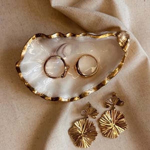 May include: A white oyster shell trinket dish with a metallic gold painted rim, containing two smooth gold-toned rings. Beside the dish are two gold-toned drop earrings, each featuring a small fan-shaped top piece and a larger, textured, circular fan-shaped pendant.
