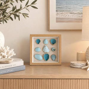 May include: A square wooden shadow box frame containing nine assorted seashells arranged in a grid. Each shell is painted in shades of blue and turquoise with gold-painted edges, mounted against a light beige fabric background within the frame.
