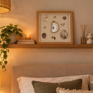 May include: A square wooden shadow box frame containing a collection of various seashells arranged on a light beige background. The display includes a large central oyster shell, smaller spiral shells, and textured bivalves in neutral tones.