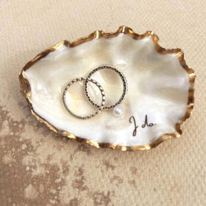 May include: A white oyster shell trinket dish with a gold-painted rim, containing two silver-toned rings and a small white pearl. The interior of the shell features the handwritten black cursive text "I do" near the bottom edge of the dish.