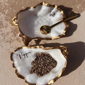 May include: Two oyster shell dishes with gold-painted edges and white interiors. One shell is labelled Salt and contains coarse white salt with a small gold spoon. The other shell is labelled Pepper and contains ground black pepper.