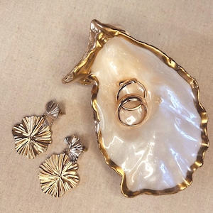May include: A decorative oyster shell dish with a pearlescent interior and a metallic gold-painted rim. Inside the shell are two plain gold-tone rings. Beside the shell are two gold-tone drop earrings featuring a textured, fan-like petal design.