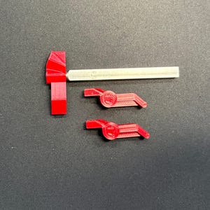 Può includere: Un componente rosso stampato in 3D a forma di T fissato a una lunga asta di metallo argentato, accompagnato da due pezzi angolari rossi più piccoli stampati in 3D. Ogni pezzo rosso presenta un centro circolare con una superficie ruvida.