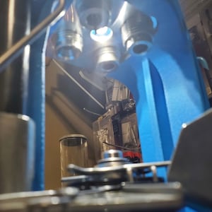 Puede incluir: Primer plano de una prensa de recarga azul con una cabeza de matriz de metal plateado. La cabeza de matriz tiene tres agujeros para diferentes matrices de recarga. La prensa está en un taller.