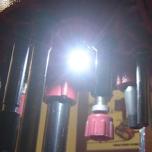 Puede incluir: Primer plano de una prensa de recarga roja con múltiples matrices de carga adjuntas. La prensa está iluminada por una fuente de luz brillante.