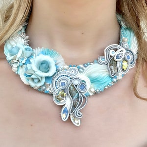 May include: A delicate, white and blue ribbon necklace with a floral design. The necklace features a large, blue flower with smaller white flowers and pearls. The necklace is made with intricate ribbon work and is a beautiful statement piece.