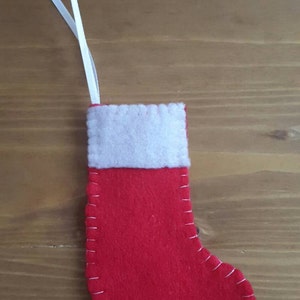 May include: A small, red felt Christmas stocking with a white cuff and a white ribbon for hanging. The stocking is stitched with white thread and is set against a wooden background.