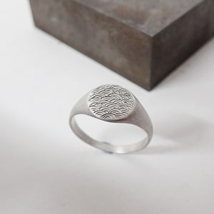 May include: A silver signet ring with a circular design featuring a pattern of overlapping waves.