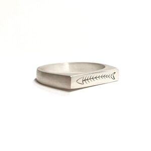 Puede incluir: Un anillo de plata con una banda rectangular y un diseño de espina de pescado grabado en la parte superior.