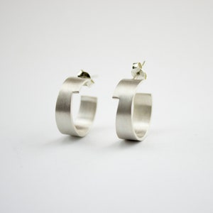 May include: A pair of silver hoop earrings with a unique, slightly asymmetrical design. The earrings are made of smooth, brushed metal and have a modern, minimalist aesthetic.