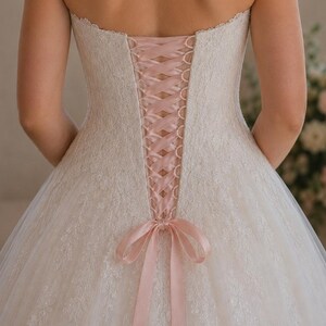 May include: A strapless white lace wedding dress with a pink ribbon corset back. The dress features a full skirt and a delicate lace pattern. The back of the dress is laced up with a pink satin ribbon, tied into a bow at the waist.