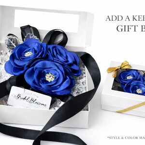 May include: Two white gift boxes containing blue fabric flowers with rhinestone centers and black ribbons. The open box includes a white tag with the text Budal Blossoms, while the closed box features a gold ribbon and a clear window.