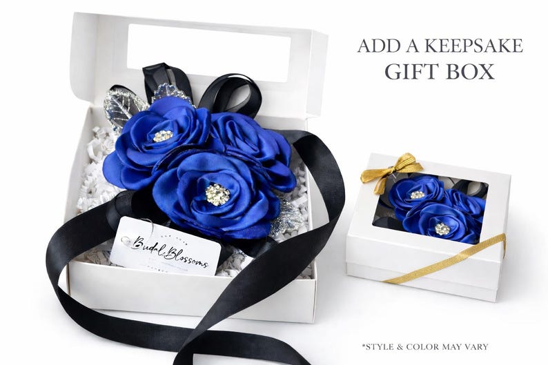 May include: Two white gift boxes containing blue fabric flowers with rhinestone centers and black ribbons. The open box includes a white tag with the text Budal Blossoms, while the closed box features a gold ribbon and a clear window.
