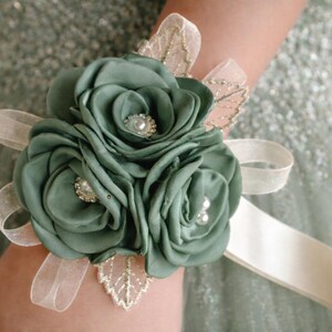May include: A wrist corsage featuring three sage green fabric roses with pearl and rhinestone centers. The floral arrangement is accented with sheer cream ribbon loops and delicate gold-trimmed leaf embellishments, secured by a wide cream satin ribbon.