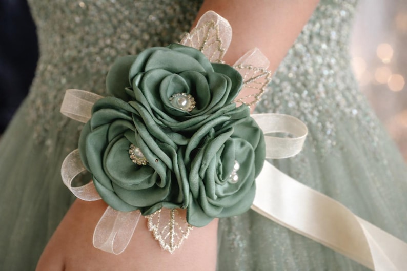 May include: A wrist corsage featuring three sage green fabric roses with pearl and rhinestone centers. The floral arrangement is accented with sheer cream ribbon loops and delicate gold-trimmed leaf embellishments, secured by a wide cream satin ribbon.
