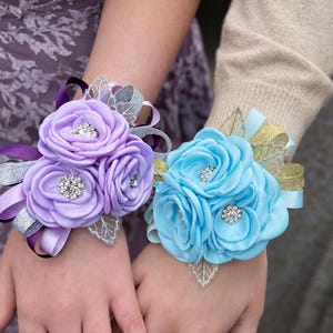 May include: Two wrist corsages featuring fabric roses in lavender and light blue. Each corsage includes silver and gold glittered ribbon accents, silver-toned leaf embellishments, and small circular rhinestone centers within the flower petals.
