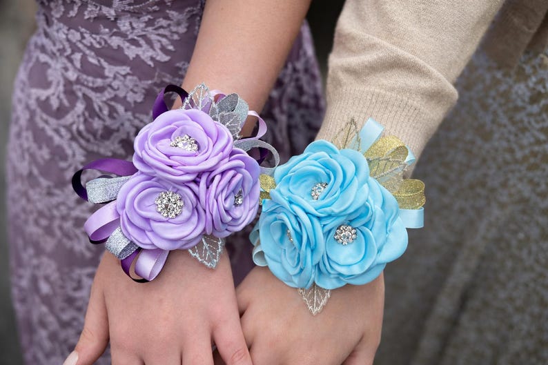 May include: Two wrist corsages featuring fabric roses in lavender and light blue. Each corsage includes silver and gold glittered ribbon accents, silver-toned leaf embellishments, and small circular rhinestone centers within the flower petals.