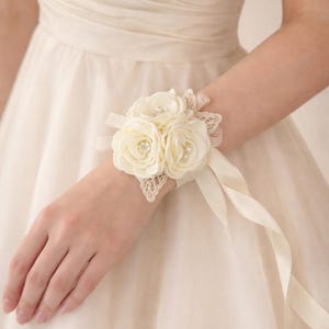 May include: A cream-colored wrist corsage featuring three fabric roses with small pearl centers, accented by delicate lace leaves and a matching satin ribbon tie. The floral arrangement is secured to a soft fabric band worn around the wrist.