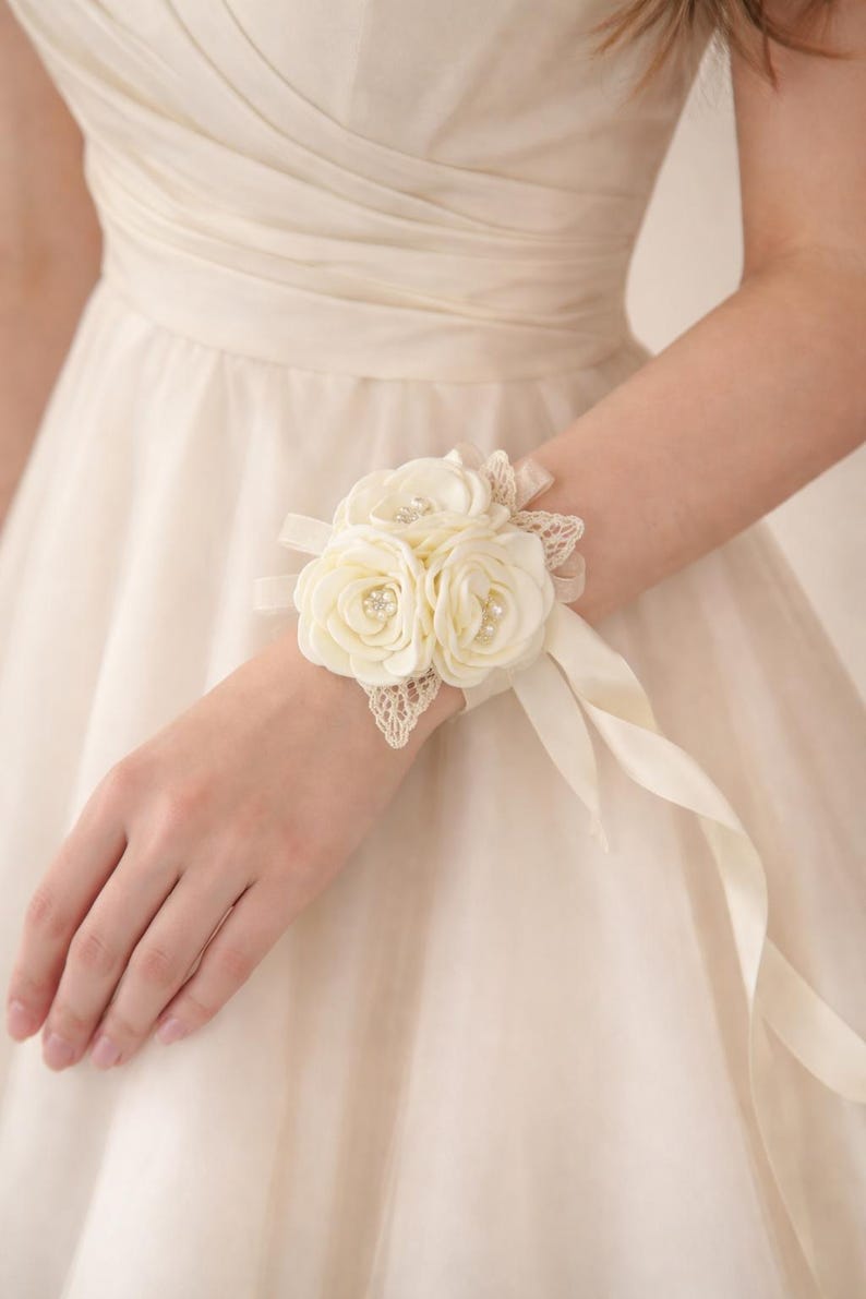 May include: A cream-colored wrist corsage featuring three fabric roses with small pearl centers, accented by delicate lace leaves and a matching satin ribbon tie. The floral arrangement is secured to a soft fabric band worn around the wrist.