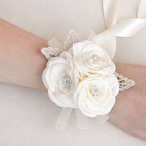 May include: A cream-colored wrist corsage featuring three fabric roses, two of which are accented with small clear rhinestones and one with a central pearl bead. The floral arrangement is decorated with delicate lace leaves and satin ribbons.