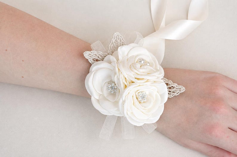 May include: A cream-colored wrist corsage featuring three fabric roses, two of which are accented with small clear rhinestones and one with a central pearl bead. The floral arrangement is decorated with delicate lace leaves and satin ribbons.
