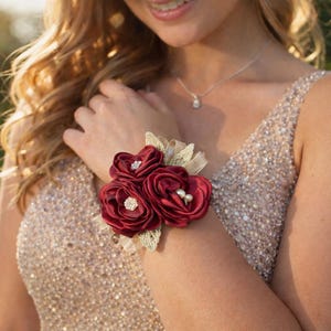 May include: A wrist corsage featuring three deep burgundy satin fabric roses. The center of two roses is accented with small clear rhinestones, while the third rose features two small white pearls. Gold-toned lace leaves sit behind the flowers.