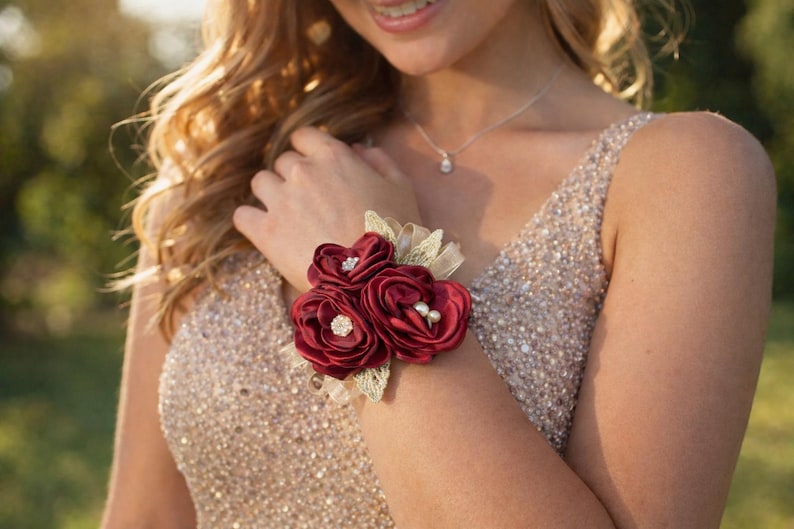 May include: A wrist corsage featuring three deep burgundy satin fabric roses. The center of two roses is accented with small clear rhinestones, while the third rose features two small white pearls. Gold-toned lace leaves sit behind the flowers.