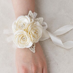 May include: A cream-colored wrist corsage featuring three satin fabric roses with small pearl and rhinestone centers. The floral arrangement is accented with delicate lace leaves and sheer ribbon ties that extend from the sides to secure the piece.