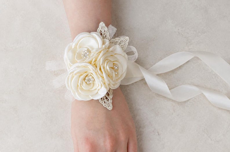 May include: A cream-colored wrist corsage featuring three satin fabric roses with small pearl and rhinestone centers. The floral arrangement is accented with delicate lace leaves and sheer ribbon ties that extend from the sides to secure the piece.