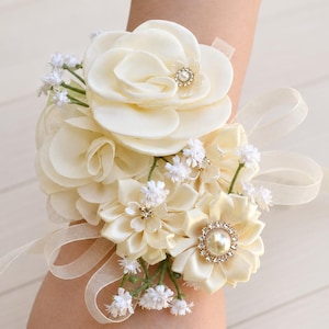 May include: A wrist corsage featuring cream-coloured satin fabric flowers, small white baby's breath accents, and clear rhinestone embellishments. The floral arrangement is secured with a sheer cream ribbon tied in a bow around the wrist.