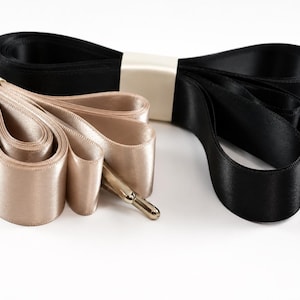 May include: Two sets of flat satin ribbon shoelaces, one in a champagne beige colour and one in black. Each set is folded and secured with a cream-coloured paper band, featuring metallic aglets at the ends of the ribbons for a finished look.