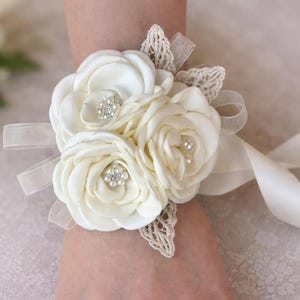 May include: A cream-colored wrist corsage featuring three fabric roses with satin petals. The center of each rose is accented with small clear rhinestones and white faux pearls. The floral arrangement is decorated with beige lace leaves and sheer ribbon ties.