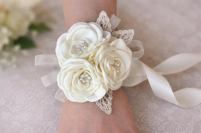 May include: A cream-colored wrist corsage featuring three fabric roses with satin petals. The center of each rose is accented with small clear rhinestones and white faux pearls. The floral arrangement is decorated with beige lace leaves and sheer ribbon ties.